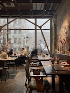 A cozy and modern cafe with large windows letting in natural light. There are people seated at tables, engaged in conversations. The interior features wooden furniture, decorative plants, and artistic elements including dried flowers and sculptures. The atmosphere is relaxed and inviting.