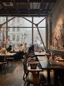 A cozy and modern cafe with large windows letting in natural light. There are people seated at tables, engaged in conversations. The interior features wooden furniture, decorative plants, and artistic elements including dried flowers and sculptures. The atmosphere is relaxed and inviting.