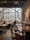 A cozy and modern cafe with large windows letting in natural light. There are people seated at tables, engaged in conversations. The interior features wooden furniture, decorative plants, and artistic elements including dried flowers and sculptures. The atmosphere is relaxed and inviting.