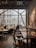 A cozy and modern cafe with large windows letting in natural light. There are people seated at tables, engaged in conversations. The interior features wooden furniture, decorative plants, and artistic elements including dried flowers and sculptures. The atmosphere is relaxed and inviting.