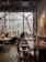 A cozy and modern cafe with large windows letting in natural light. There are people seated at tables, engaged in conversations. The interior features wooden furniture, decorative plants, and artistic elements including dried flowers and sculptures. The atmosphere is relaxed and inviting.