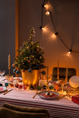 A festive table setting features a small Christmas tree in a golden pot adorned with lights and baubles. The table is covered with a red and white striped cloth and is set with plates, napkins, and cutlery. Warm lighting creates a cozy atmosphere, and string lights add a decorative touch.