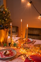 A warm, inviting holiday table setting decorated with golden candles, a small Christmas tree in a gold pot adorned with baubles, and lush fairy lights. The table is covered with a red and white striped tablecloth, complete with neatly set plates featuring red napkins with snowflake patterns, wine glasses, and a small dessert on each plate.