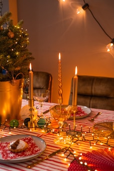 A warm, inviting holiday table setting decorated with golden candles, a small Christmas tree in a gold pot adorned with baubles, and lush fairy lights. The table is covered with a red and white striped tablecloth, complete with neatly set plates featuring red napkins with snowflake patterns, wine glasses, and a small dessert on each plate.