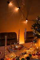 A festively decorated table with a striped tablecloth features elegant candle holders with tall, slender candles. The table is adorned with string lights and assorted baubles, adding a warm, inviting glow. A small decorated tree is visible to one side, and the background is enhanced with softly lit string lights casting gentle shadows.