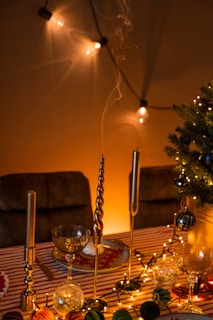 A festively decorated table with a striped tablecloth features elegant candle holders with tall, slender candles. The table is adorned with string lights and assorted baubles, adding a warm, inviting glow. A small decorated tree is visible to one side, and the background is enhanced with softly lit string lights casting gentle shadows.