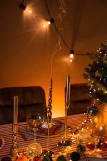 A festively decorated table with a striped tablecloth features elegant candle holders with tall, slender candles. The table is adorned with string lights and assorted baubles, adding a warm, inviting glow. A small decorated tree is visible to one side, and the background is enhanced with softly lit string lights casting gentle shadows.