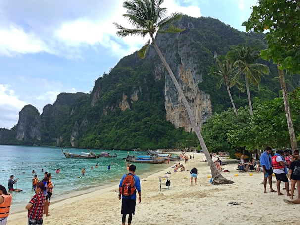 A bustling tropical beach scene with people swimming and relaxing on the sand. Long-tail boats are anchored near the shore, and lush green cliffs rise dramatically in the background. Palm trees sway in the breeze, adding to the idyllic setting.