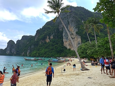 A bustling tropical beach scene with people swimming and relaxing on the sand. Long-tail boats are anchored near the shore, and lush green cliffs rise dramatically in the background. Palm trees sway in the breeze, adding to the idyllic setting.