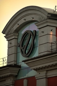 An ornate architectural facade with classical elements features a prominent large circular emblem that appears embossed in black. The structure includes decorative cornices and archways, positioned against a glass backdrop under an early evening sky.
