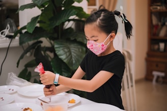 A young girl wearing a decorative face mask and a black shirt is engaged in an activity at a table. She is holding a red icing bag, possibly decorating or preparing food, with several plates around her. The setting is indoors, with a large green plant and some wooden furniture visible in the background.