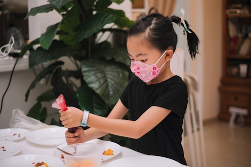 A young girl wearing a decorative face mask and a black shirt is engaged in an activity at a table. She is holding a red icing bag, possibly decorating or preparing food, with several plates around her. The setting is indoors, with a large green plant and some wooden furniture visible in the background.