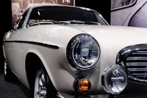 A classic vintage car displayed in a showroom with a cream-colored body and shiny chrome detailing. The front of the car features prominent circular headlights, an orange indicator light, and a sleek grille. Reflected light on the car surface emphasizes its smooth finish.