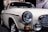 A classic vintage car displayed in a showroom with a cream-colored body and shiny chrome detailing. The front of the car features prominent circular headlights, an orange indicator light, and a sleek grille. Reflected light on the car surface emphasizes its smooth finish.