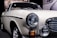 A classic vintage car displayed in a showroom with a cream-colored body and shiny chrome detailing. The front of the car features prominent circular headlights, an orange indicator light, and a sleek grille. Reflected light on the car surface emphasizes its smooth finish.