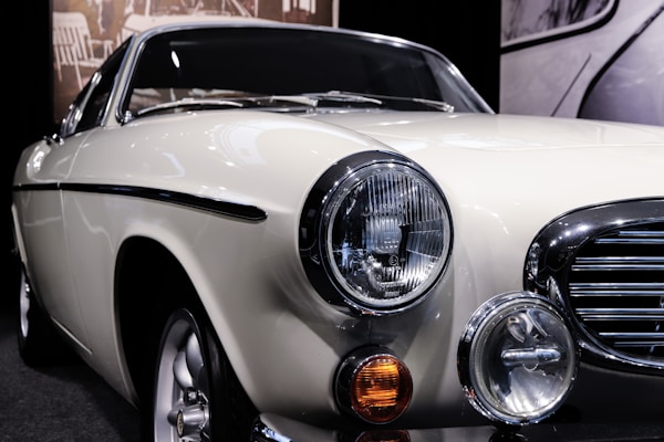 A classic vintage car displayed in a showroom with a cream-colored body and shiny chrome detailing. The front of the car features prominent circular headlights, an orange indicator light, and a sleek grille. Reflected light on the car surface emphasizes its smooth finish.
