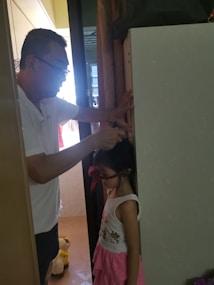 A man is marking the height of a young girl on a wall or a door frame. The girl is wearing a pink skirt and a white tank top with a cute design. There is a stuffed animal partially visible on the floor behind them. The setting is a dimly lit room, and the man is wearing glasses and a casual white shirt.