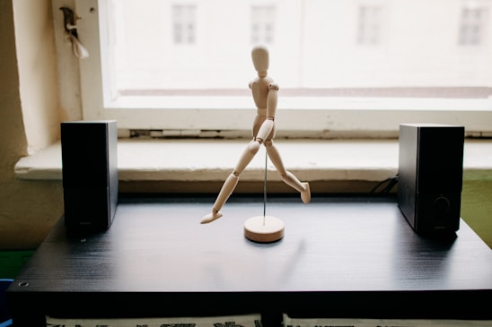 A wooden mannequin is positioned in a dynamic, running pose on a round stand. It is placed on a dark wooden table with two black rectangular speakers on either side. The background shows a bright window, letting in natural light.