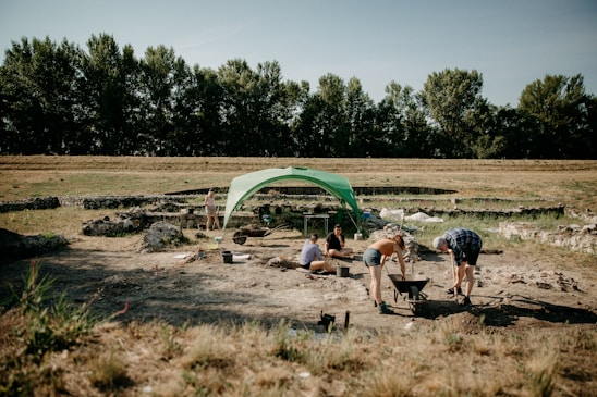 A group of people is participating in an archaeological excavation under a green canopy tent. The setting appears to be an open field with scattered stone ruins and tools. Trees line the background under a clear sky.