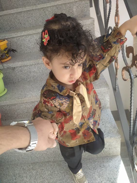 A young child with curly hair, adorned with hair accessories, is holding an adult's hand while standing on a staircase. The child is wearing a colorful floral-patterned shirt and looking sideways. One hand is placed on the staircase railing, and they appear to be stepping upwards.