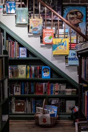 A cozy, well-organized bookstore corner with books displayed on shelves and steps. Colorful children's books are propped up on the stairs, and there is a basket labeled 'Keep the Kids Busy Books' filled with additional titles. The wooden floors and bookshelves give a warm and inviting atmosphere.