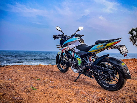 A motorcycle is parked on a rocky cliff overlooking the ocean. The bike has vibrant decals and a sleek design, set against a backdrop of a cloudy sky and gentle waves. A small palm tree is visible to the right.
