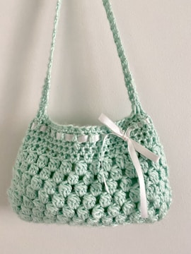 A small, handmade crochet bag with a textured pattern and a single strap. The bag features a decorative white ribbon tied into a bow on the front.