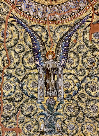 A detailed mosaic artwork depicting an angel with outspread wings, adorned with intricate patterns and vibrant colors. The background is filled with swirling floral motifs and geometric shapes, creating a rich tapestry-like effect. The angel is dressed in a decorated robe, and there is a sense of symmetry and elegance throughout the design.