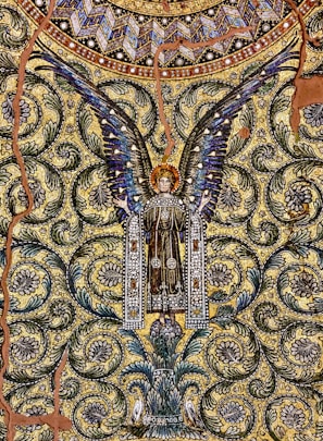 A detailed mosaic artwork depicting an angel with outspread wings, adorned with intricate patterns and vibrant colors. The background is filled with swirling floral motifs and geometric shapes, creating a rich tapestry-like effect. The angel is dressed in a decorated robe, and there is a sense of symmetry and elegance throughout the design.