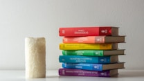 A stack of six hardcover books in various colors is neatly arranged on a light-colored surface against a plain wall. The colors of the book covers range from red, orange, yellow, green, blue, to purple, creating a rainbow pattern. To the left of the stack, there is a textured, white cylindrical candle.