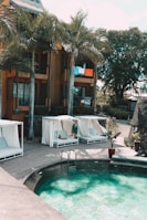 A serene poolside setting with cabanas featuring white curtains and cushions is complemented by a small pool with clear blue-green water. A person in a robe is sitting outside one of the cabanas, and palm trees provide a tropical ambiance. The background includes a multi-story building with brightly colored towels hanging over the balcony railings.