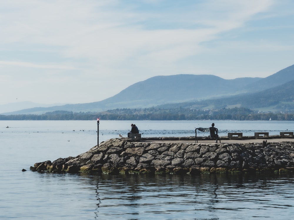 a couple people sitting on a rock wall by a body of water