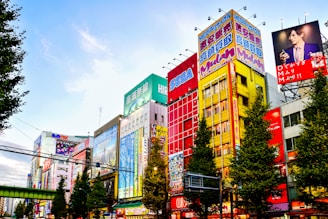 Colorful buildings adorned with large, vibrant billboards and advertisements in a bustling urban setting. The scene is animated by a mix of bright colors and diverse ads, reflecting a lively city atmosphere. Trees lined along the street add a touch of greenery.