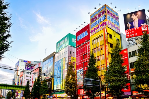 Colorful buildings adorned with large, vibrant billboards and advertisements in a bustling urban setting. The scene is animated by a mix of bright colors and diverse ads, reflecting a lively city atmosphere. Trees lined along the street add a touch of greenery.