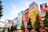 Colorful buildings adorned with large, vibrant billboards and advertisements in a bustling urban setting. The scene is animated by a mix of bright colors and diverse ads, reflecting a lively city atmosphere. Trees lined along the street add a touch of greenery.