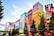 Colorful buildings adorned with large, vibrant billboards and advertisements in a bustling urban setting. The scene is animated by a mix of bright colors and diverse ads, reflecting a lively city atmosphere. Trees lined along the street add a touch of greenery.