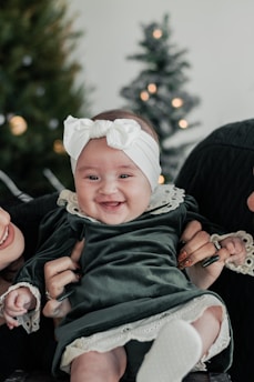 A baby dressed in a green velvet dress with white lace trim and wearing a white headband with a bow is being held by two adults. The baby is smiling joyfully. There are Christmas trees in the background with blurry lights that add to the festive ambiance.