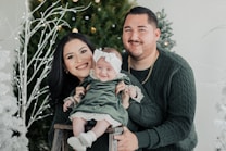 A smiling family poses in front of festive decorations with a green theme. The people are wearing matching dark green outfits, and they are surrounded by Christmas trees with glowing lights. The baby is dressed in a green dress with a bow on the head, sitting on a small wooden stool held by the parents.