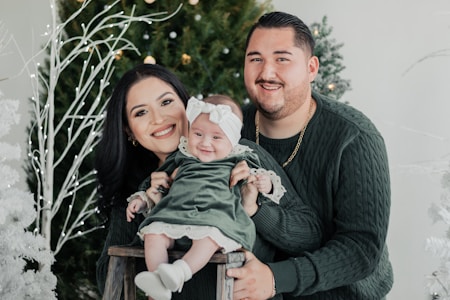 A smiling family poses in front of festive decorations with a green theme. The people are wearing matching dark green outfits, and they are surrounded by Christmas trees with glowing lights. The baby is dressed in a green dress with a bow on the head, sitting on a small wooden stool held by the parents.