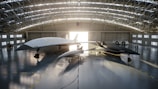 A spacious aircraft hangar houses two sleek, futuristic-looking jet aircraft. The larger aircraft is white, while the smaller one is dark gray. Both aircraft have pointed noses and streamlined designs. Sunlight streams through the open hangar doors, casting long shadows on the polished concrete floor. An American flag hangs prominently at the back of the hangar, and two people are visible near a white vehicle parked to the side.