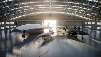 A spacious aircraft hangar houses two sleek, futuristic-looking jet aircraft. The larger aircraft is white, while the smaller one is dark gray. Both aircraft have pointed noses and streamlined designs. Sunlight streams through the open hangar doors, casting long shadows on the polished concrete floor. An American flag hangs prominently at the back of the hangar, and two people are visible near a white vehicle parked to the side.