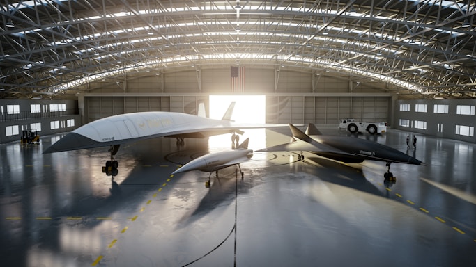 A spacious aircraft hangar houses two sleek, futuristic-looking jet aircraft. The larger aircraft is white, while the smaller one is dark gray. Both aircraft have pointed noses and streamlined designs. Sunlight streams through the open hangar doors, casting long shadows on the polished concrete floor. An American flag hangs prominently at the back of the hangar, and two people are visible near a white vehicle parked to the side.