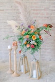 An elegant floral arrangement featuring vibrant orange, purple, and pink flowers with lush green foliage. Tall pampas grass adds a soft, wispy texture, and the arrangement is accompanied by candles placed in wooden holders and glass lanterns. The scene is set against a light-colored brick wall, creating a rustic yet refined atmosphere.
