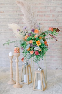 An elegant floral arrangement featuring vibrant orange, purple, and pink flowers with lush green foliage. Tall pampas grass adds a soft, wispy texture, and the arrangement is accompanied by candles placed in wooden holders and glass lanterns. The scene is set against a light-colored brick wall, creating a rustic yet refined atmosphere.