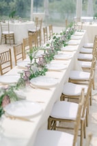 A long dining table elegantly set with white plates, golden cutlery, and decorated with a lush green and purple floral garland. The table is surrounded by wooden chairs with white cushions, situated in a bright, airy environment likely intended for an event or celebration.