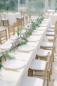 A long dining table elegantly set with white plates, golden cutlery, and decorated with a lush green and purple floral garland. The table is surrounded by wooden chairs with white cushions, situated in a bright, airy environment likely intended for an event or celebration.