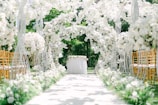 An aisle decorated with lush white flowers arching over, creating a romantic and elegant setting. Golden chairs are arranged on each side, and a table adorned with a white tablecloth and flowers is positioned at the end. The backdrop features vibrant green foliage, providing a serene, natural atmosphere.