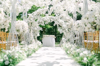 An aisle decorated with lush white flowers arching over, creating a romantic and elegant setting. Golden chairs are arranged on each side, and a table adorned with a white tablecloth and flowers is positioned at the end. The backdrop features vibrant green foliage, providing a serene, natural atmosphere.