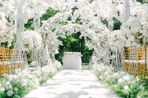 An aisle decorated with lush white flowers arching over, creating a romantic and elegant setting. Golden chairs are arranged on each side, and a table adorned with a white tablecloth and flowers is positioned at the end. The backdrop features vibrant green foliage, providing a serene, natural atmosphere.