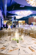 A lavishly decorated banquet hall features multiple round tables adorned with elegant table settings, including white plates, glasses, and beige napkins. The tables are centered with cylindrical glass vases filled with decorative lights and surrounded by greenery. The ambiance is enhanced by large, ornate chandeliers hanging from the ceiling, casting a warm glow across the room. Chairs with a wooden finish are arranged neatly around the tables, and the background features a softly lit atmosphere with blurred figures and decor.